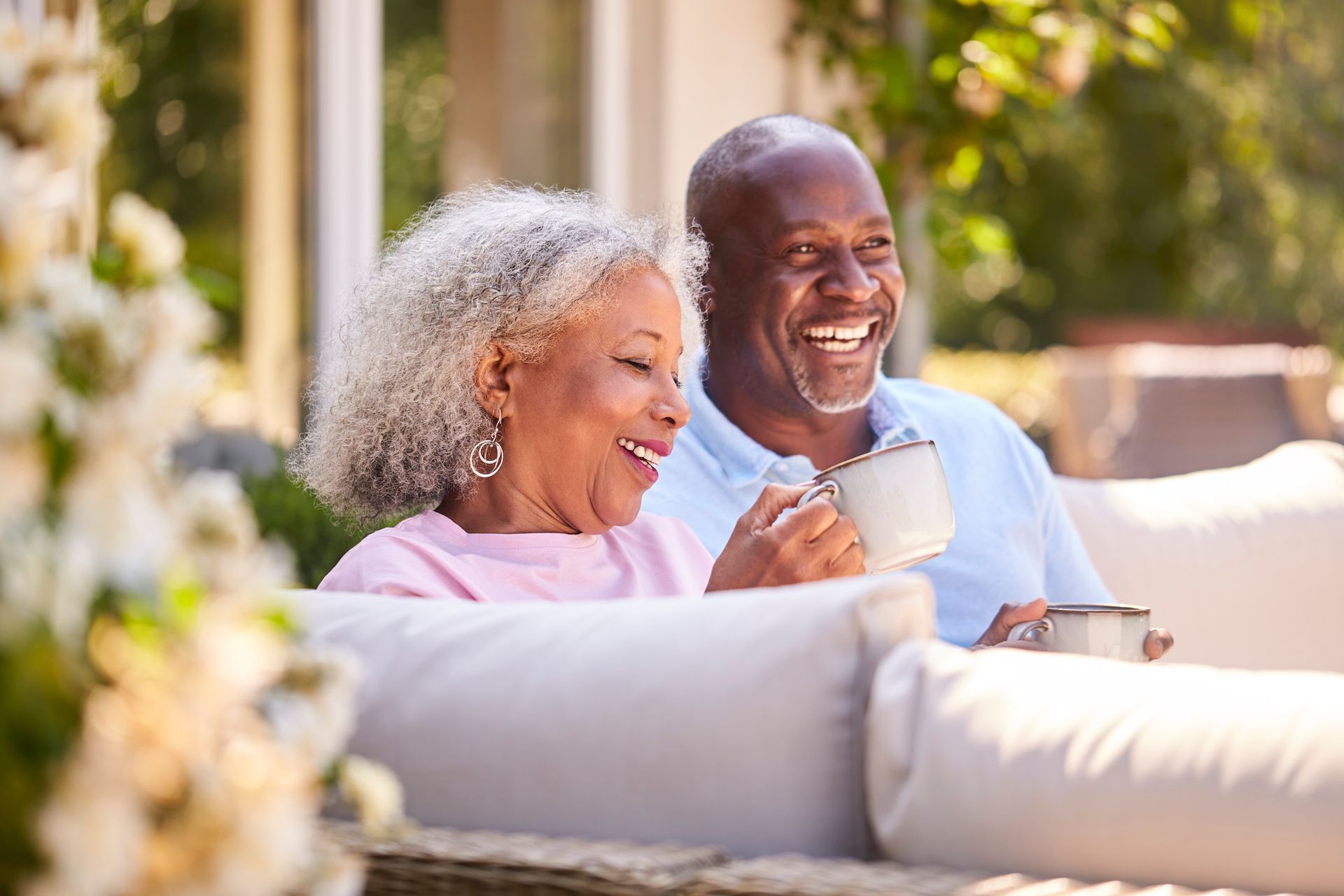 Smiling couple outdoors, holding mugs, sitting on cushions. Sunny day, white flowers in foreground.