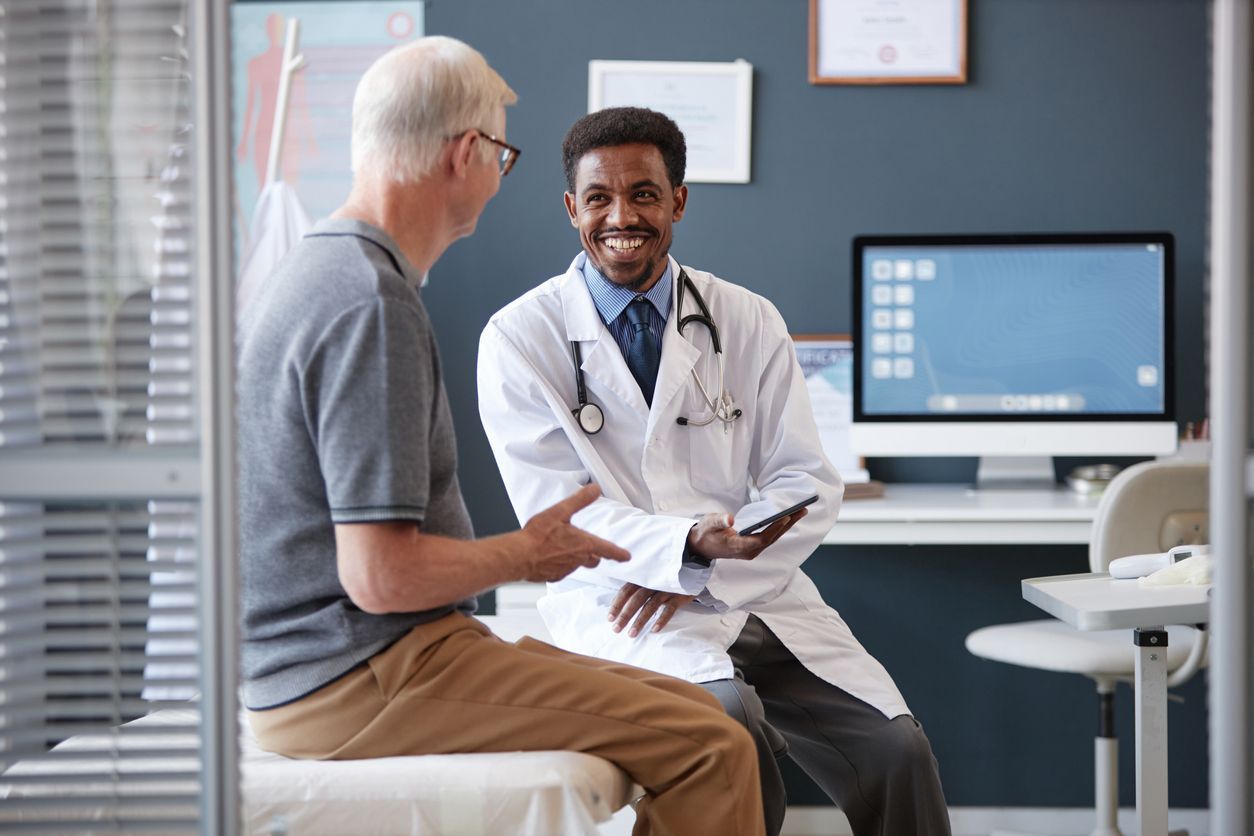 Doctor in white coat smiling, talking with a patient in a medical examination room.