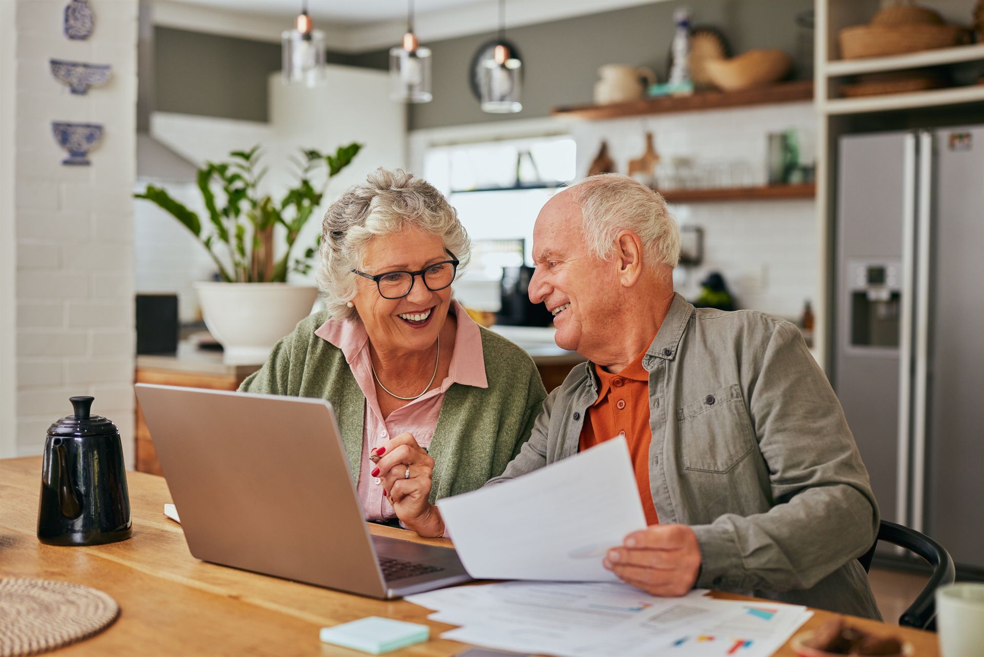Elderly couple smiling at laptop, examining documents at a kitchen table.