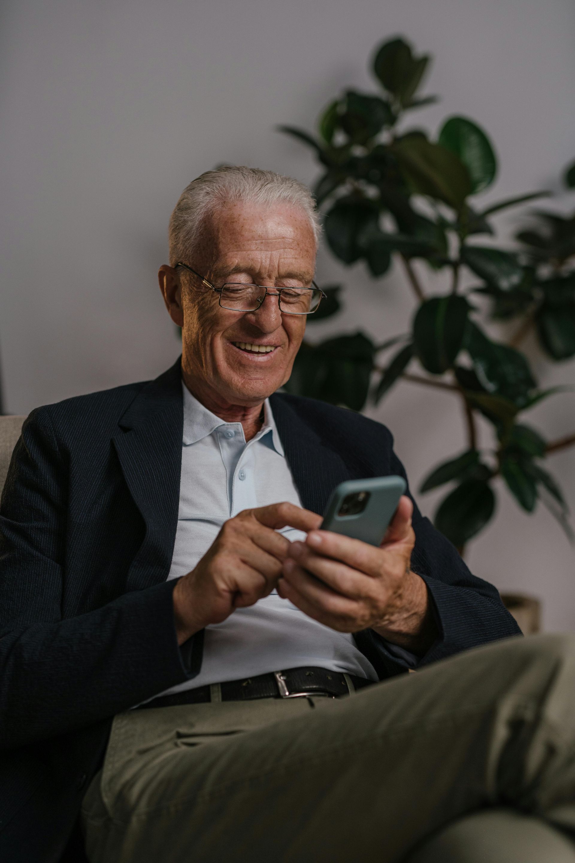 Smiling person in glasses using a smartphone; seated indoors near a plant.