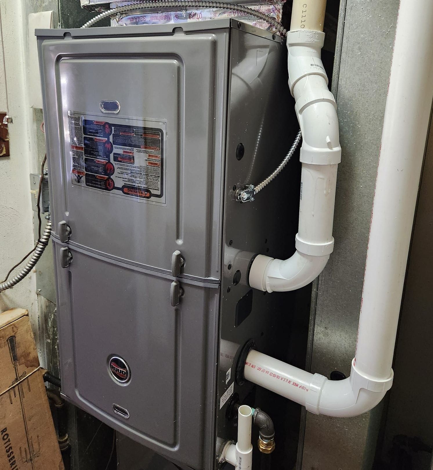 A gray furnace is sitting in a basement next to white pipes.