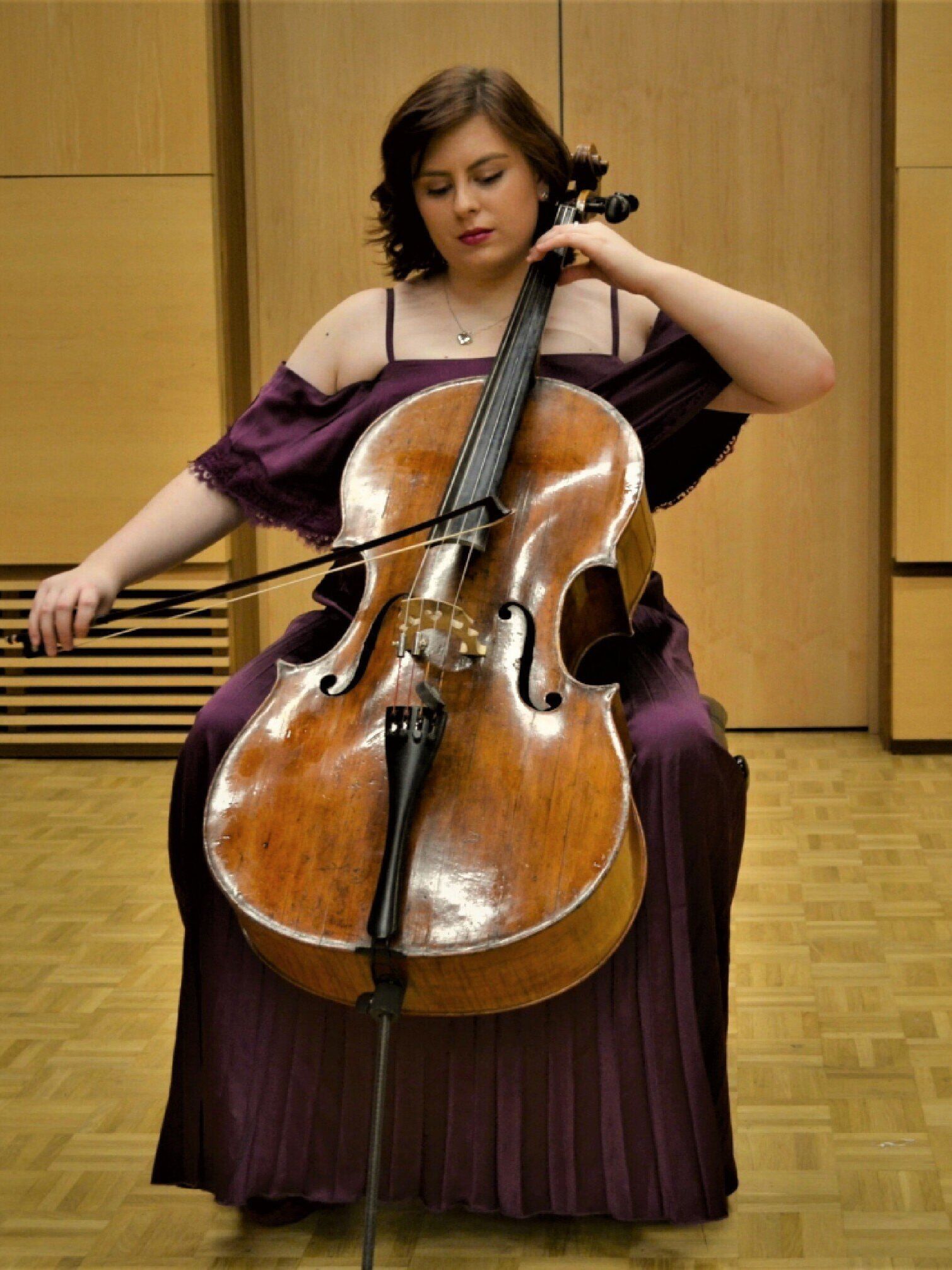 Soloist Katharine Ley, a Cello player sat on a stool in a purple off-the-shoulder dress & shoulder-length brown hair.