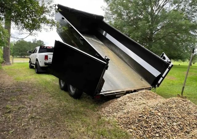 White truck with a black dump trailer unloading gravel onto a dirt patch in a grassy area.