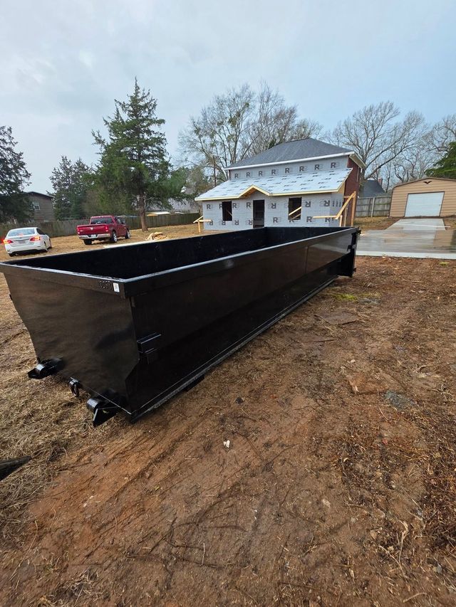 Black dumpster on a dirt lot near a house under construction. Cloudy sky in the background.