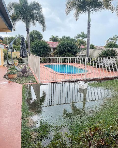 Flooded backyard with a swimming pool behind a white fence, reflecting in standing water. Overcast day.
