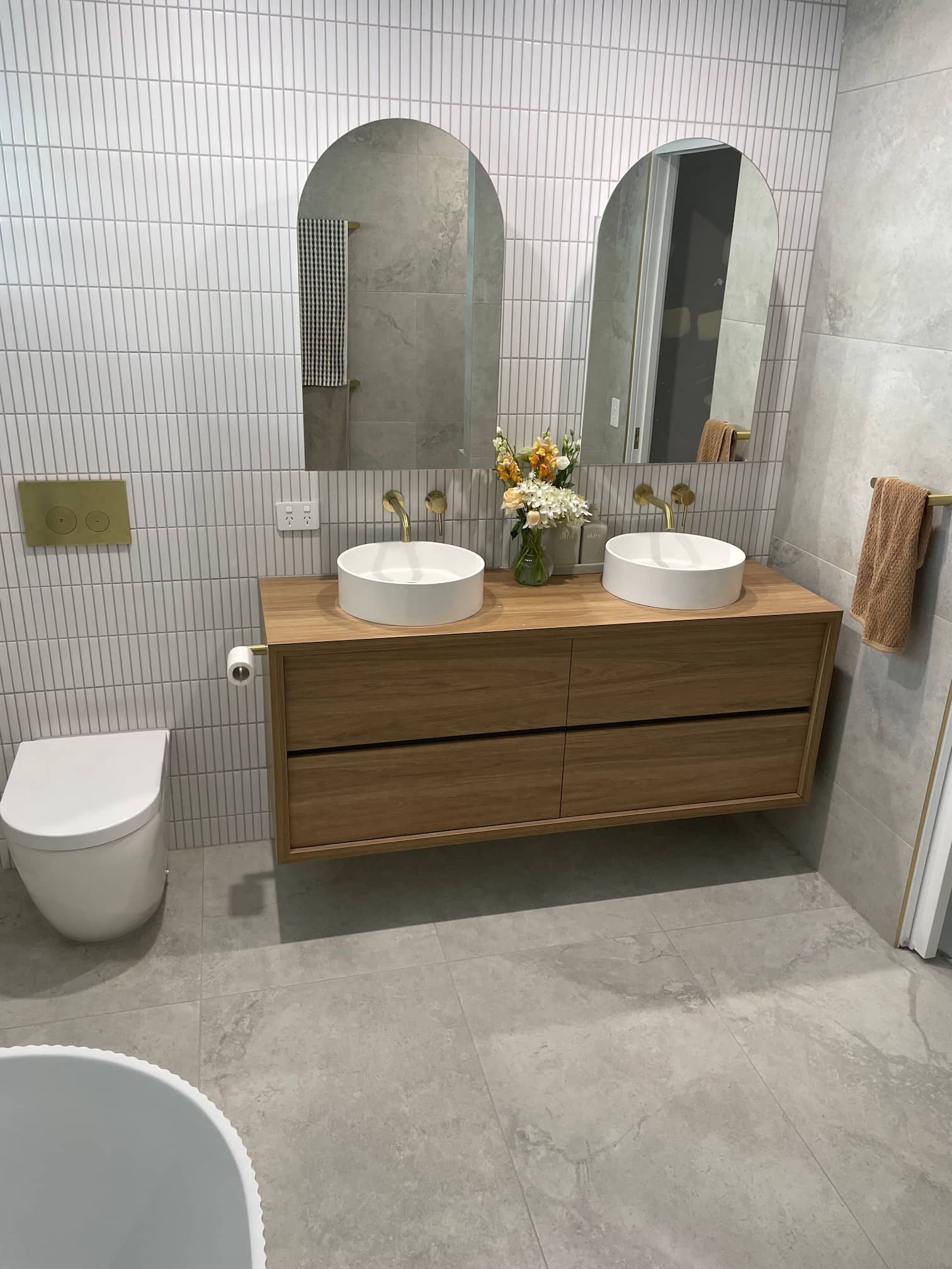 Modern bathroom with floating wooden vanity, two round sinks, arched mirrors, and patterned white tile.