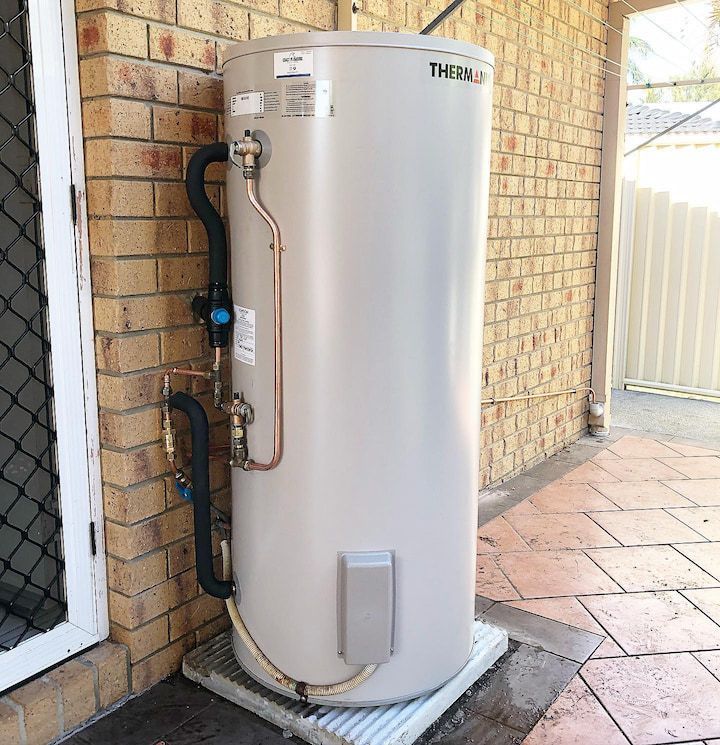 A tall, cylindrical hot water heater with copper pipes is mounted outside on a concrete pad next to a brick wall.