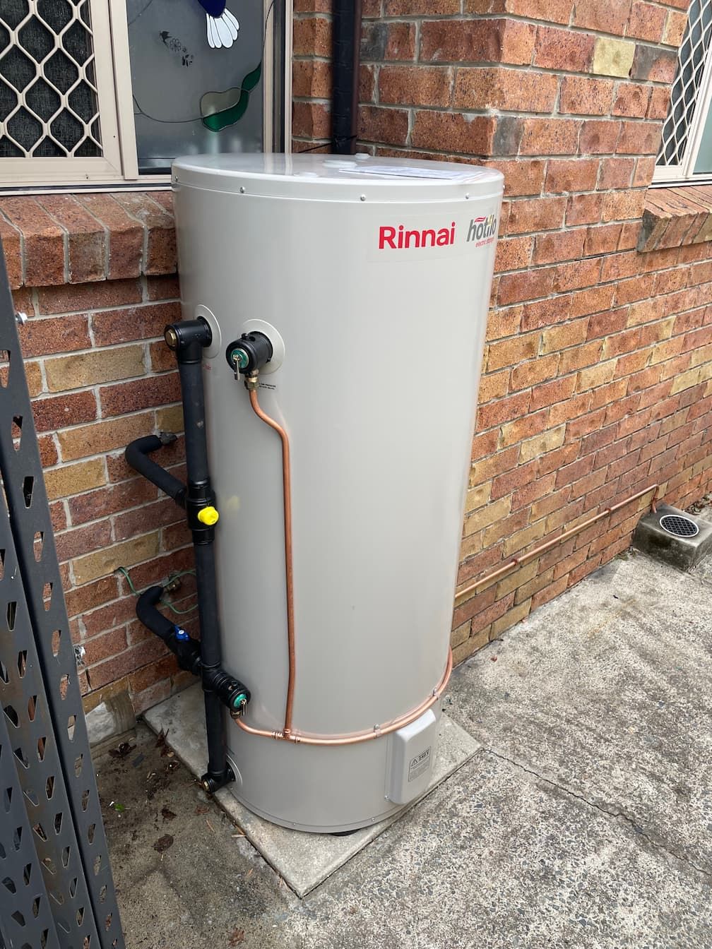 Rinnai hot water heater outdoors against a brick wall. Gray cylinder with black and copper pipes.
