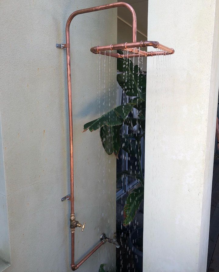 Copper outdoor shower with water cascading, mounted on a light-colored wall, with greenery behind.