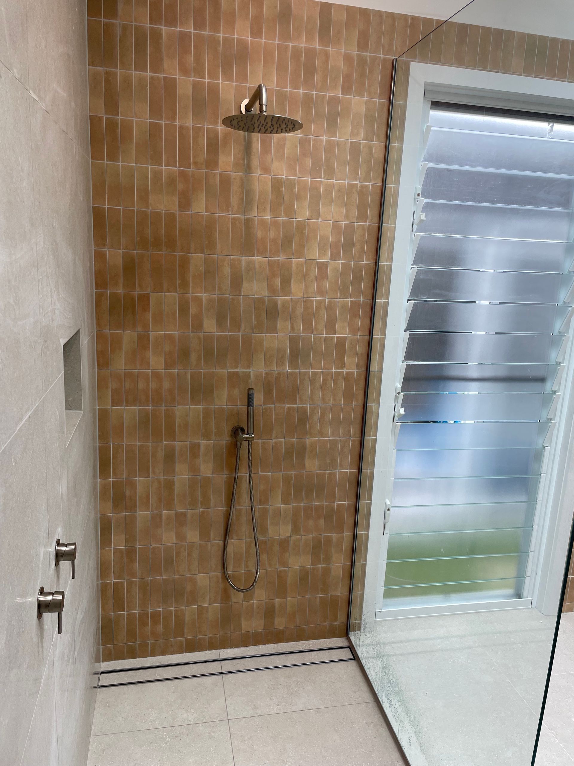 Shower with tan tile walls, handheld showerhead, open window with louvers, and a niche.