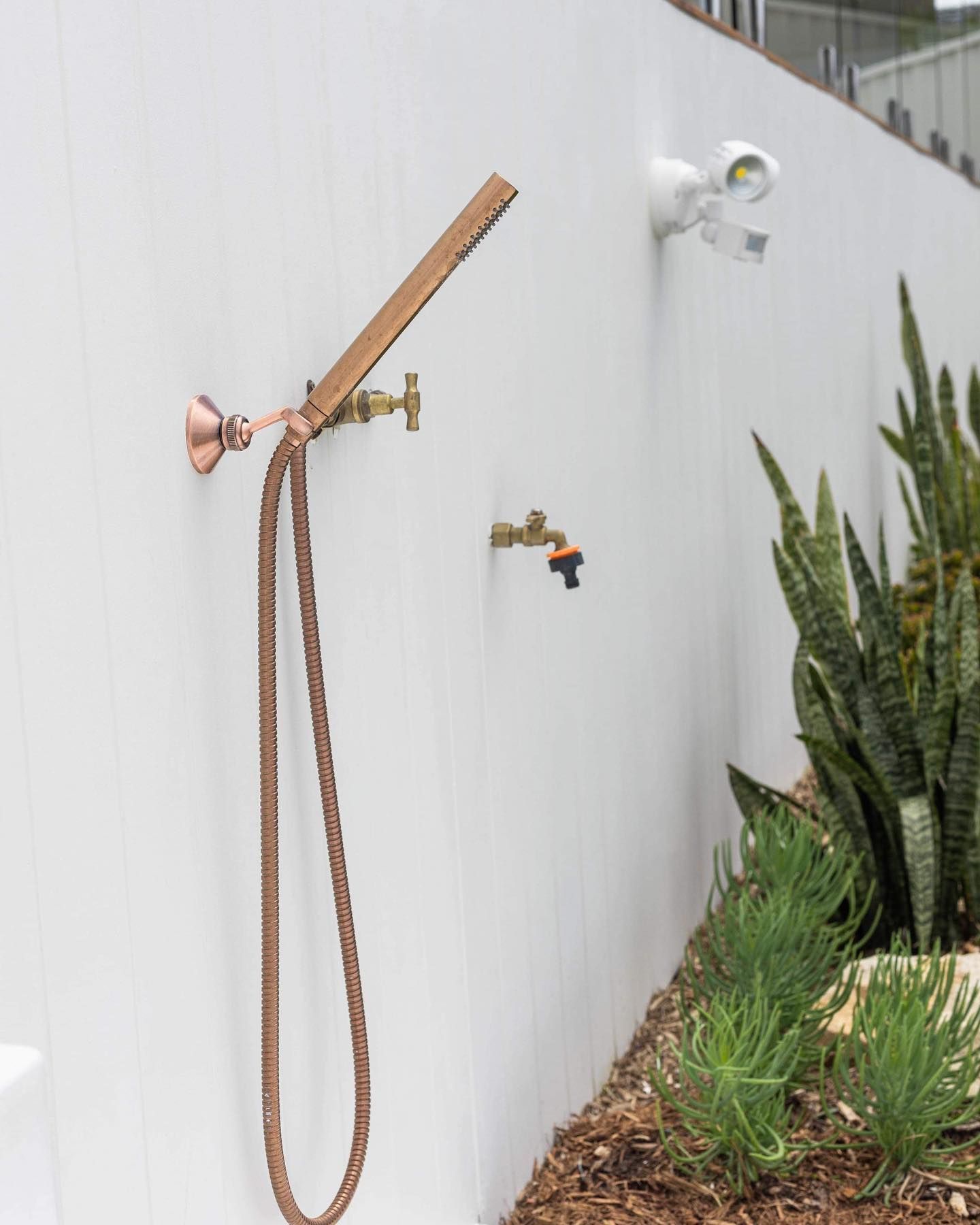 Copper outdoor shower and faucet on a white wall; plants in front. Security light above.