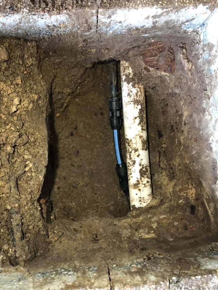 A rectangular hole in the ground reveals a vertical white pipe, connected to a black fitting and blue pipe.