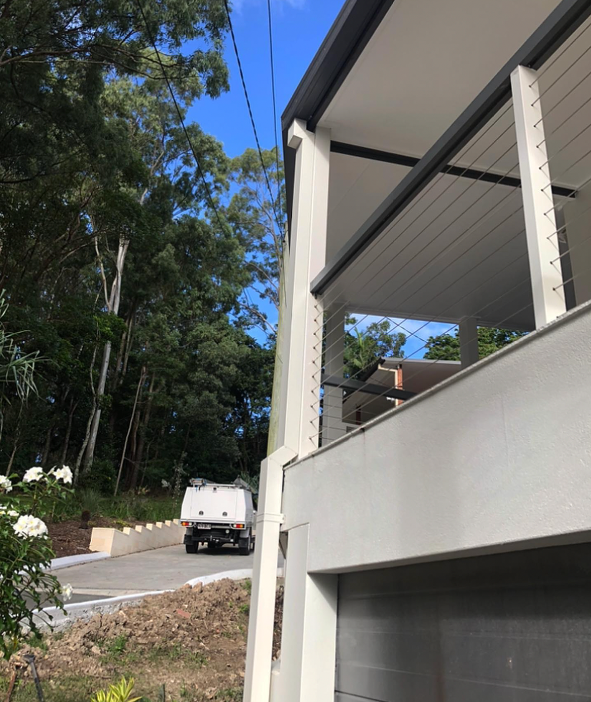 White truck on a road near a modern white house with a balcony and power lines, trees in the background.
