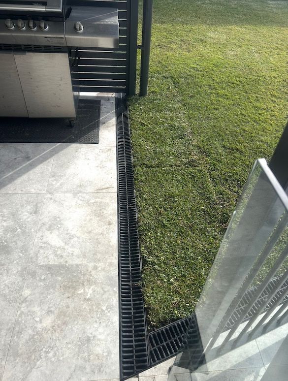 Black drain on concrete patio next to grass and a BBQ grill.