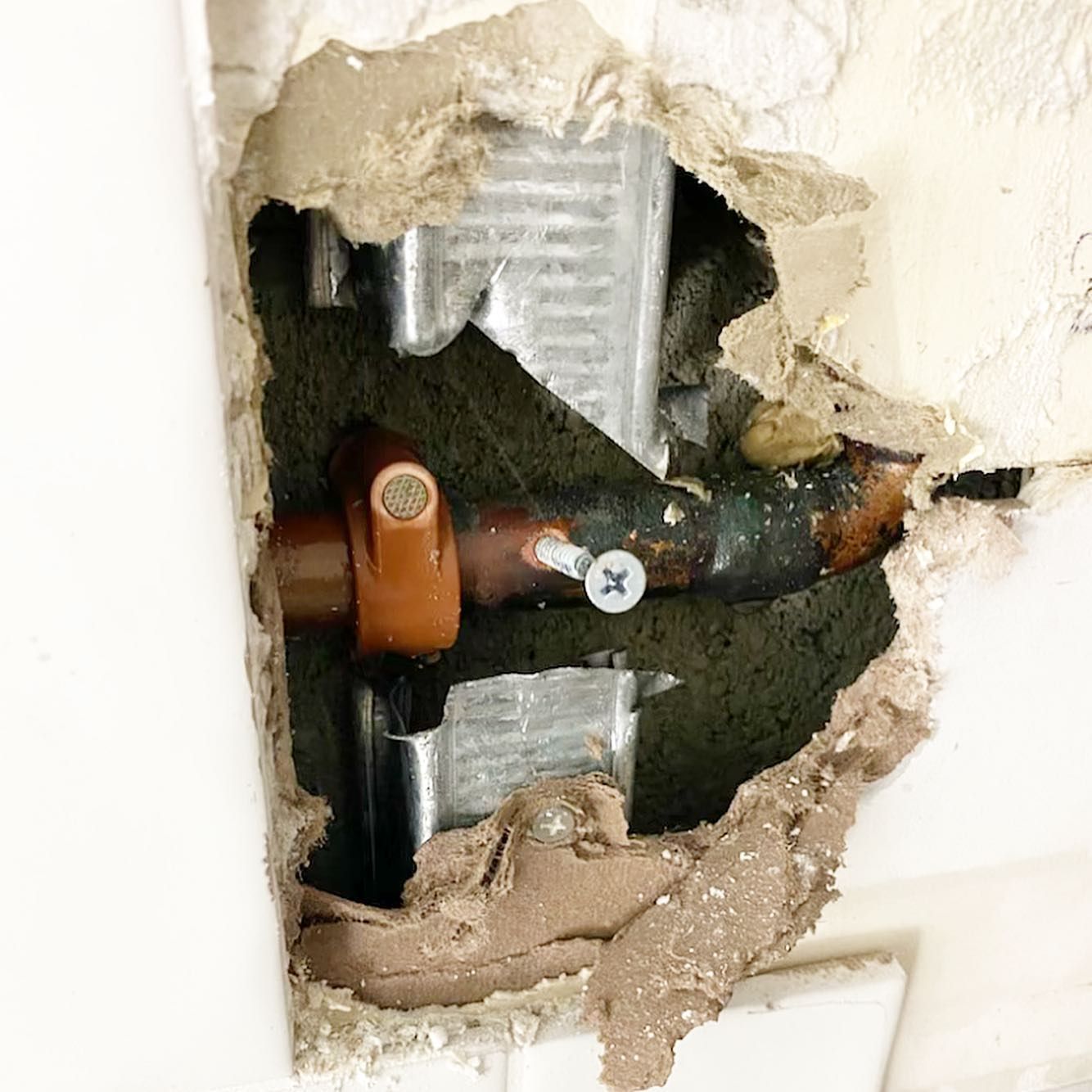 Hole in wall reveals copper pipe with valve, screw, and metal support.