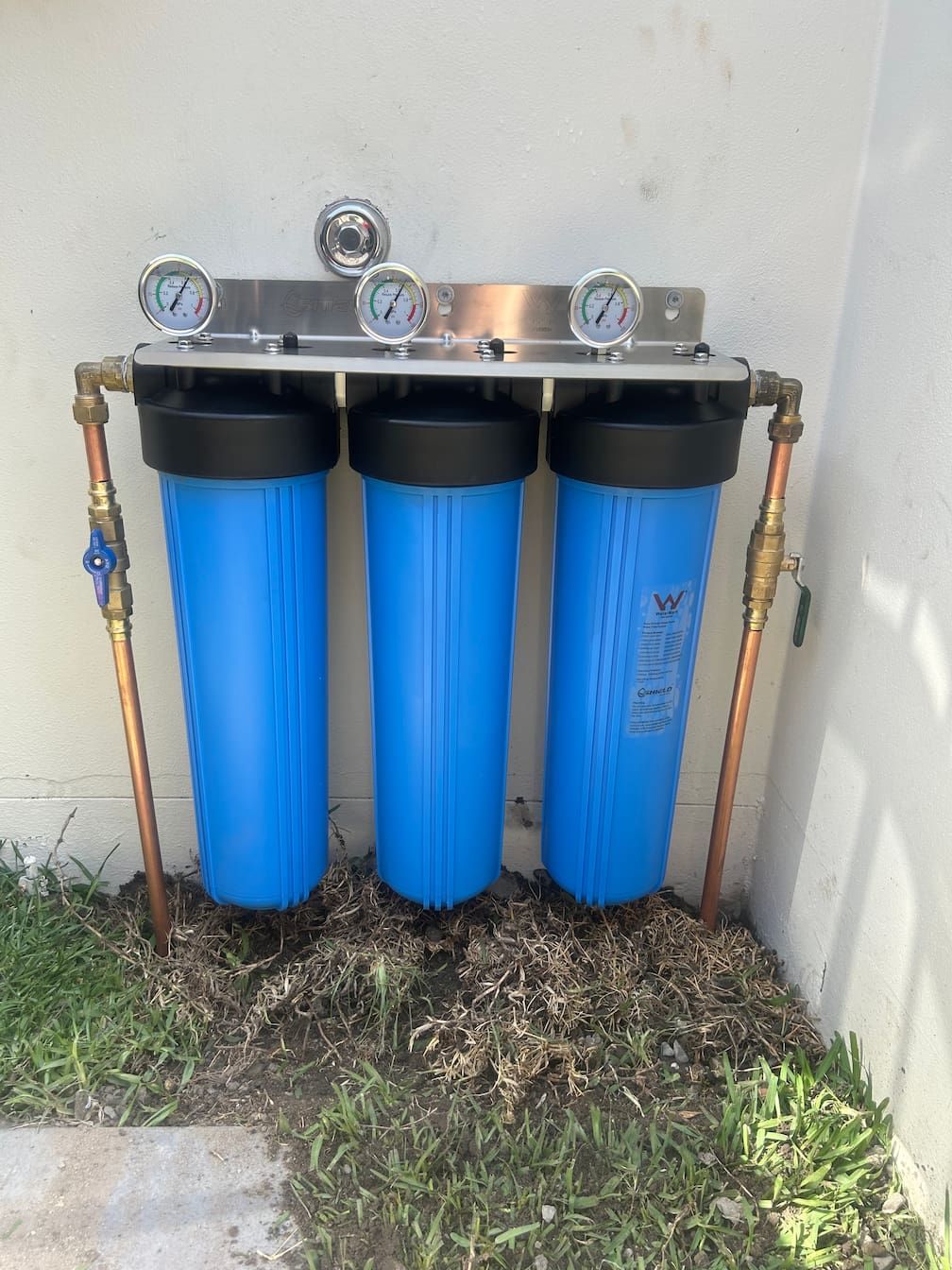 Water filtration system with three blue filter housings, copper pipes, and pressure gauges, mounted outdoors.