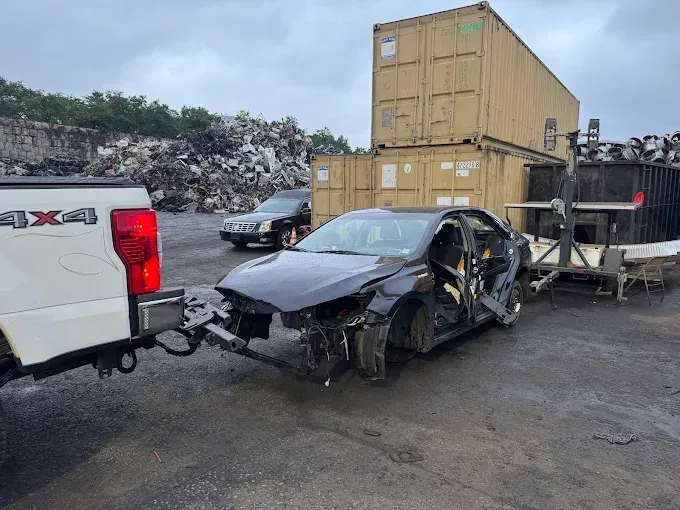 A white truck is towing a wrecked car in a scrap yard.