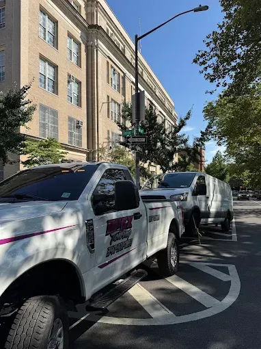 White work trucks parked on a city street in front of a multi-story beige building. Sunny day.