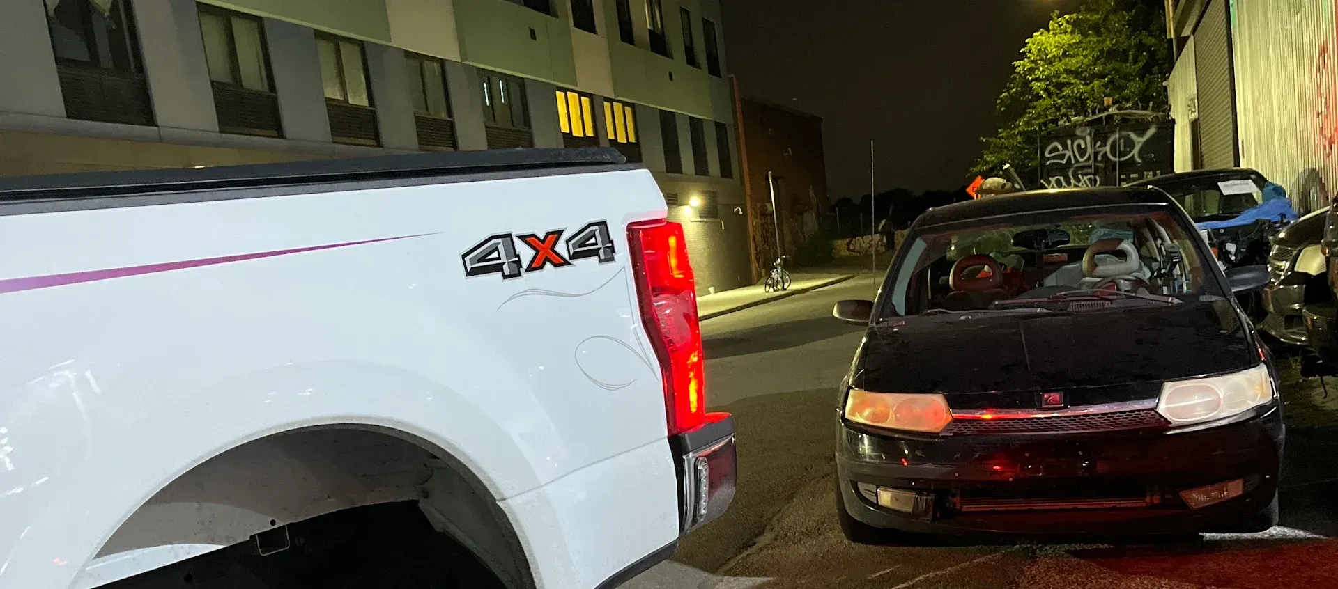 White pickup truck facing a black car on a dark street. Buildings and night lighting in the background.