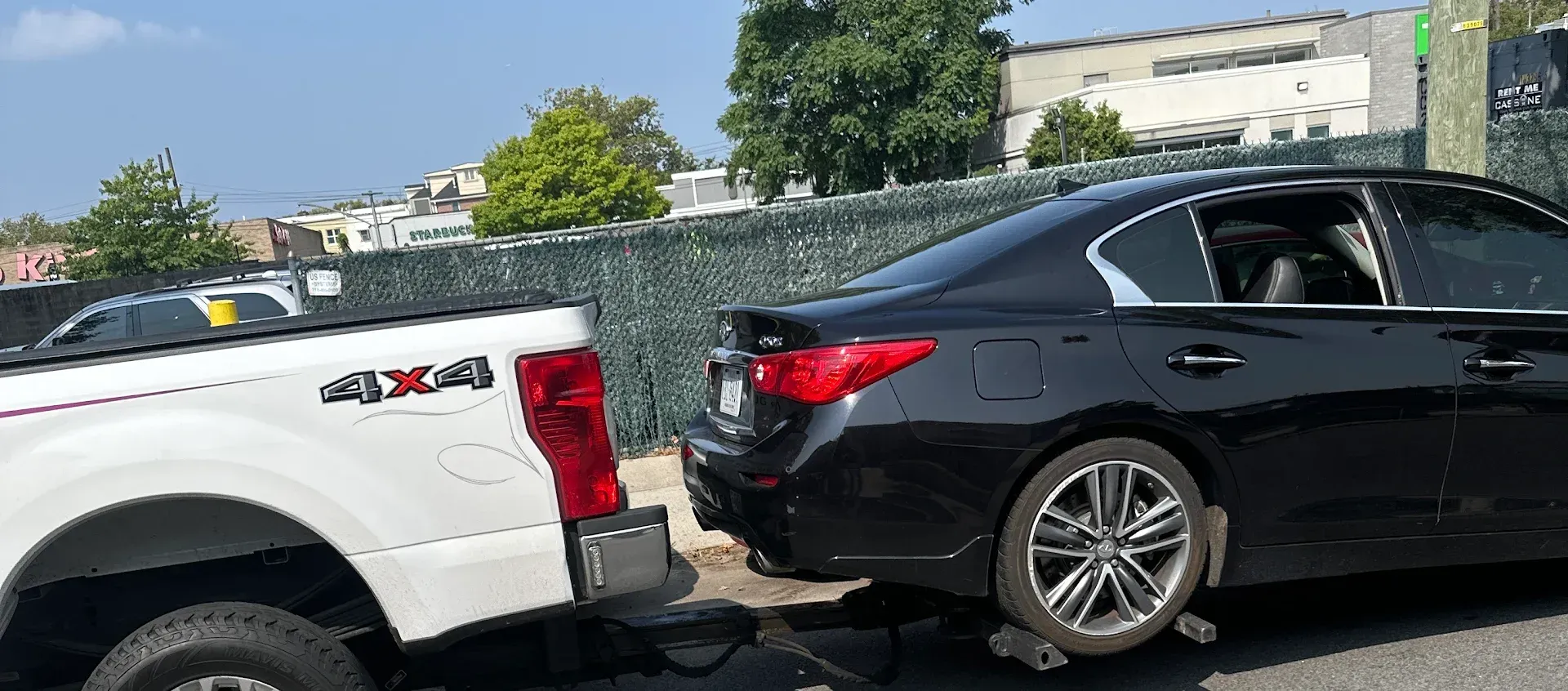 A white pickup truck towing a black car on a sunny day. The car's rear wheel is on a dolly.