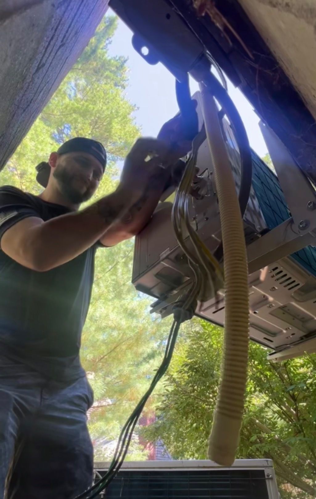 A person wearing a camo hat and work clothes installs wiring on an outdoor Carrier air conditioning unit.