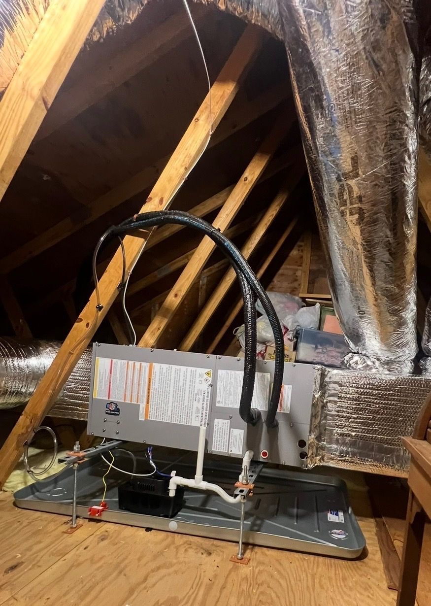 An HVAC air handler unit sits on a metal drain pan in an attic, connected to insulated silver air ducts and PVC piping.
