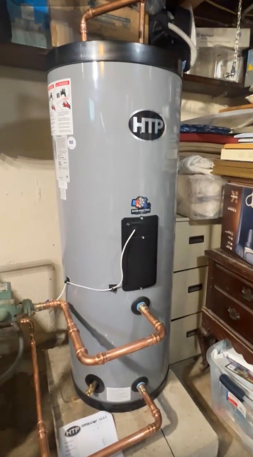 A tall, cylindrical gray HTP water heater with copper plumbing connections, installed in a cluttered indoor setting.