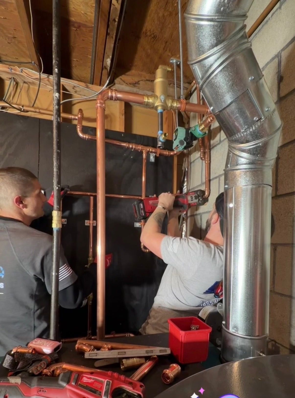 Two technicians install copper plumbing pipes near a large metal exhaust vent in a utility room.
