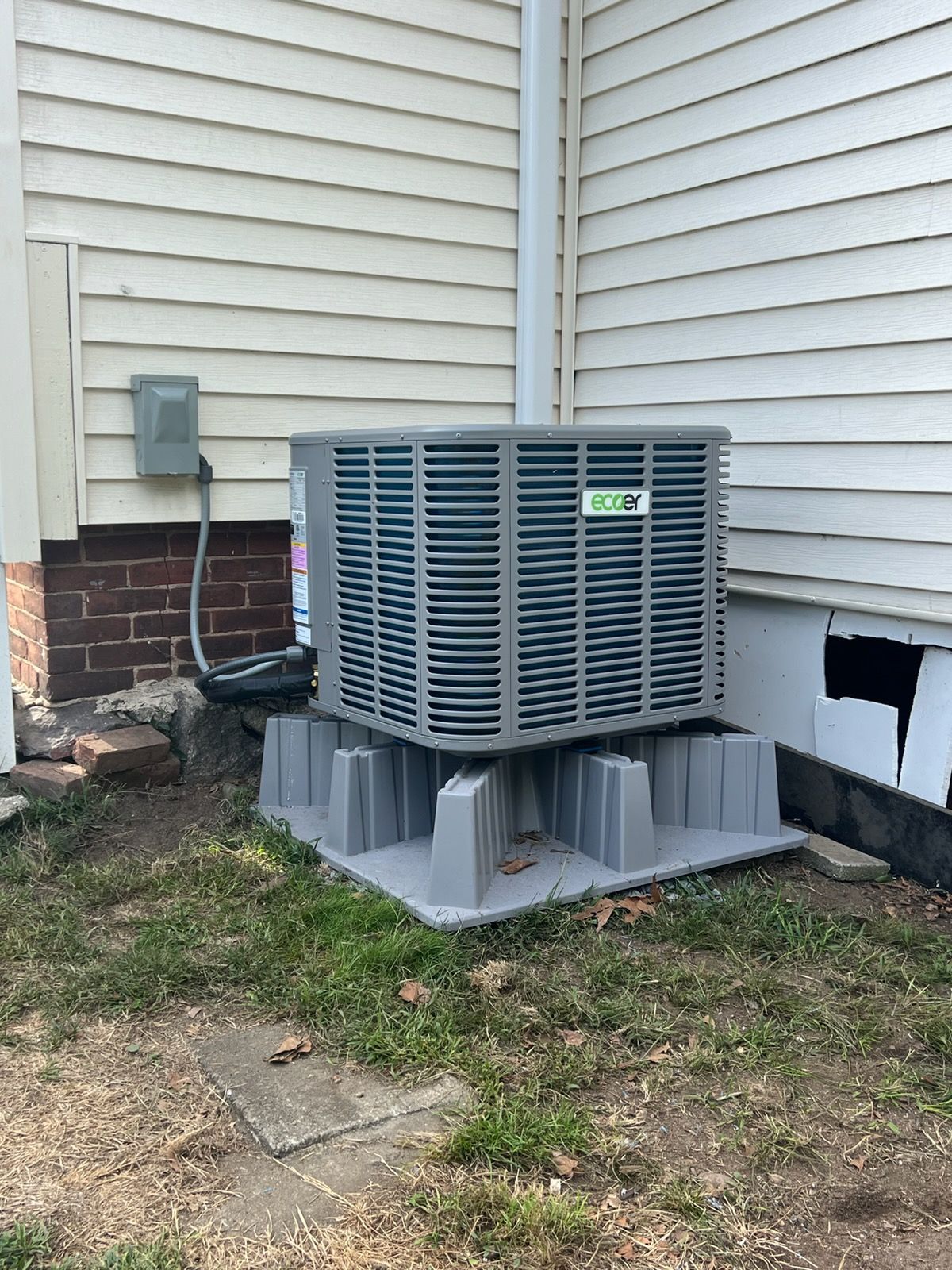 An outdoor HVAC condenser unit sits on a plastic pad next to a house with beige siding and a brick foundation.