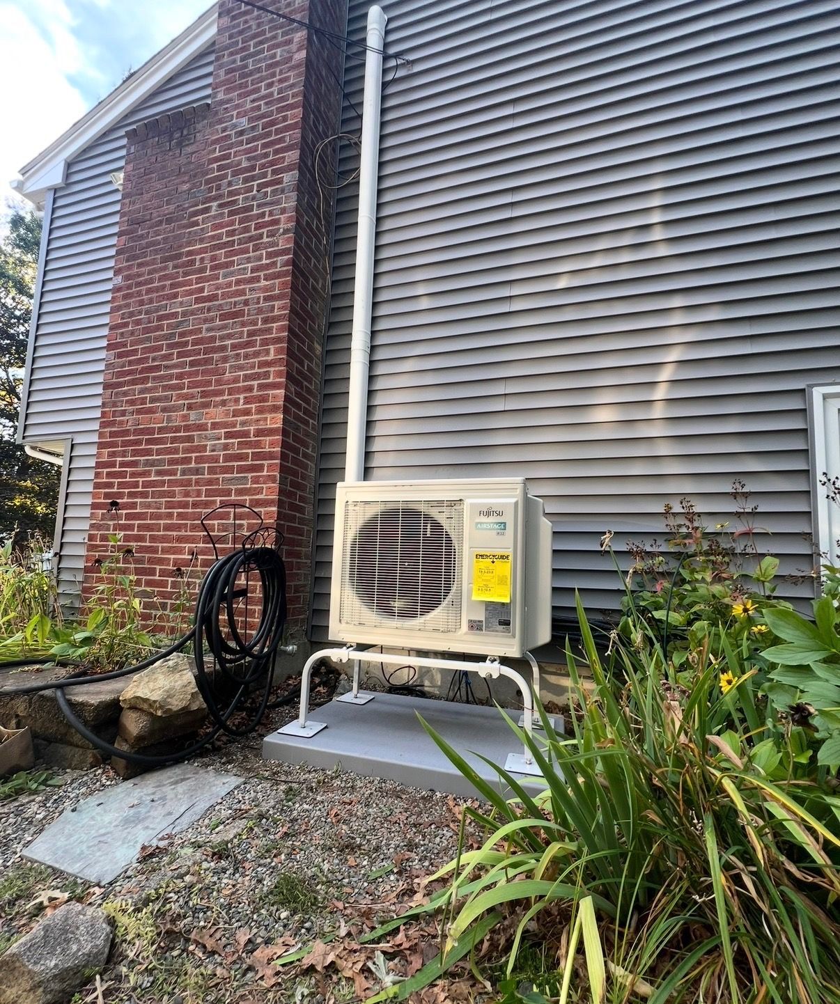 An outdoor air conditioning heat pump unit mounted on a stand next to a brick chimney and gray siding on a house.