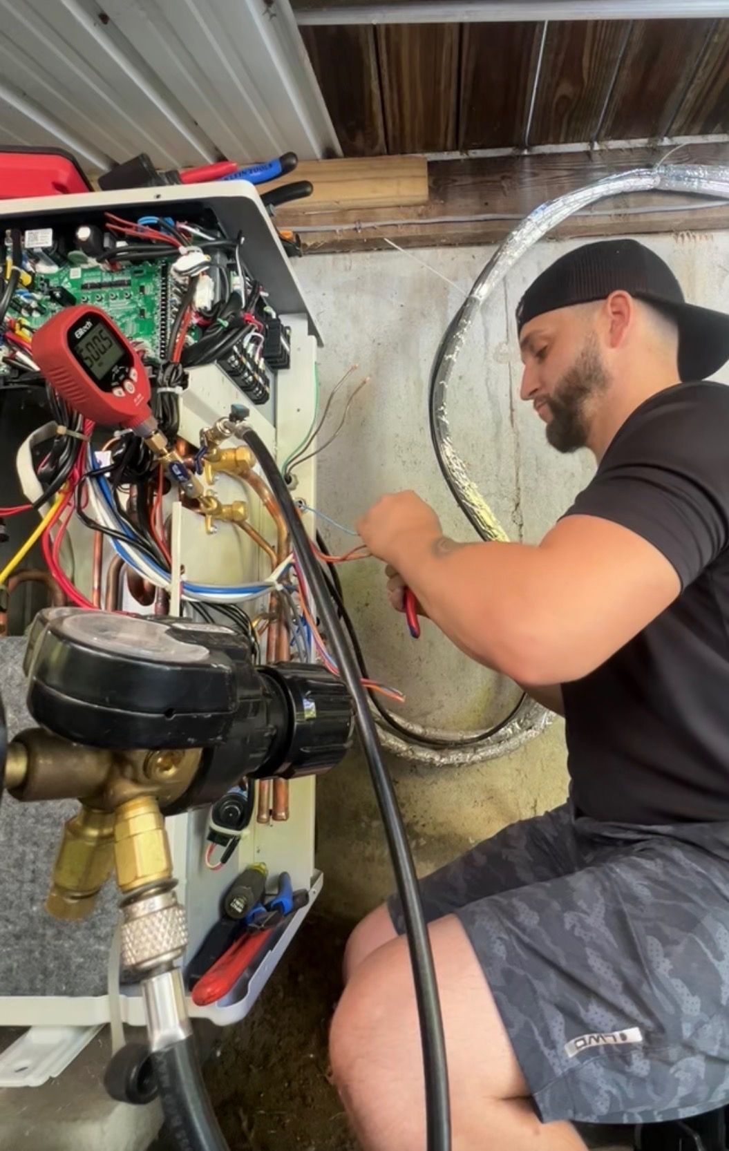 A technician works on an open HVAC system with pressure gauges, cutting a wire inside a utility room.