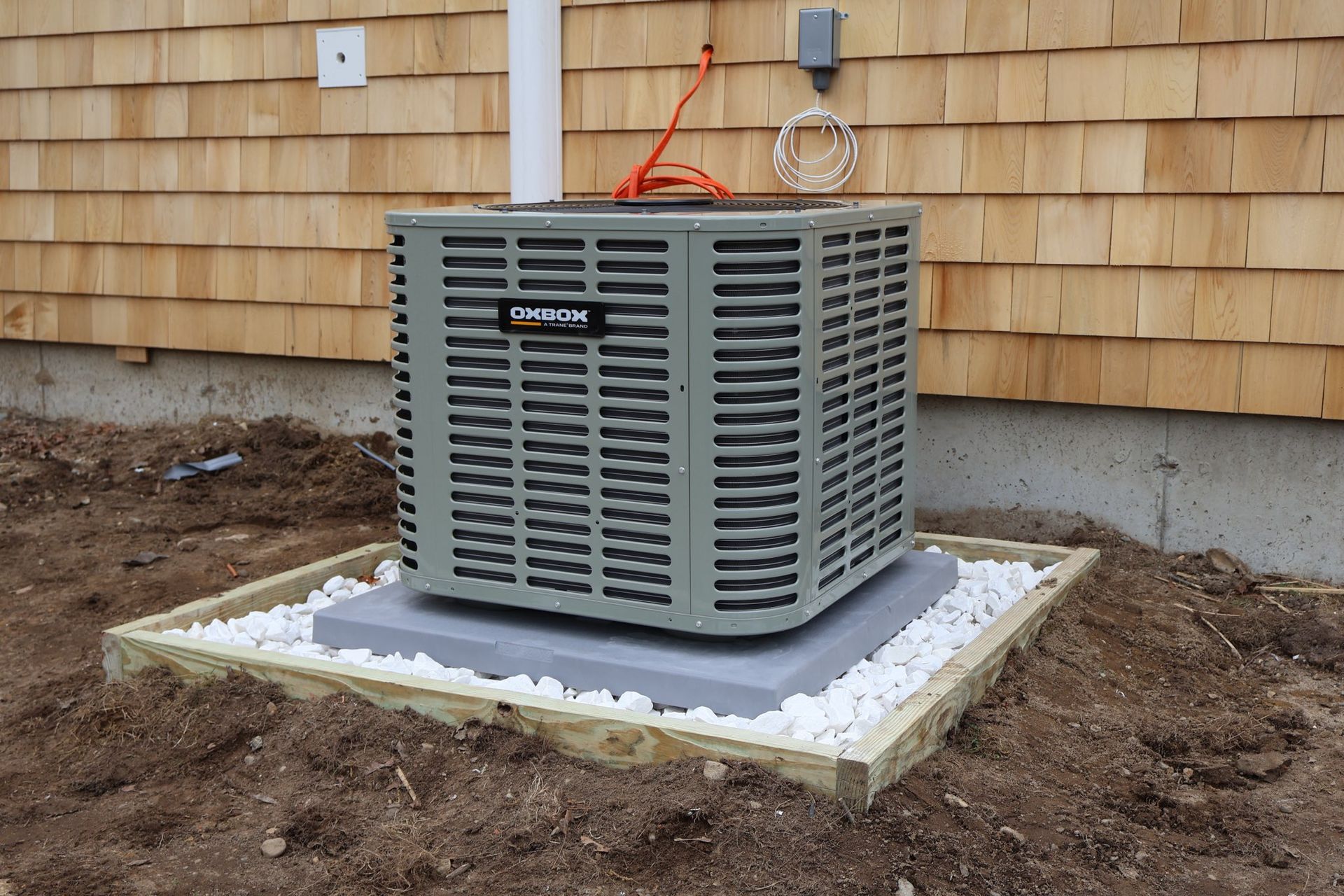 A grey central air conditioning unit sits on a concrete pad surrounded by white gravel, placed against a wood-sided house.