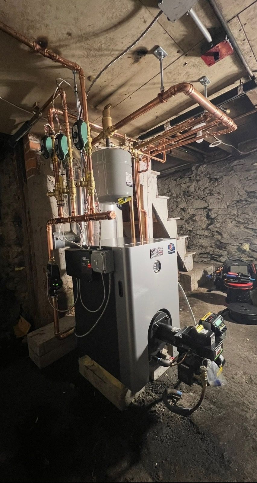 A new heating boiler with copper piping and circulator pumps installed in a basement with stone walls.
