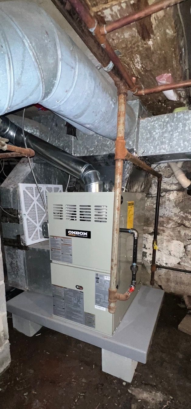 A modern, light-gray gas furnace sits on a raised platform in a rustic basement, connected to overhead metal ductwork.