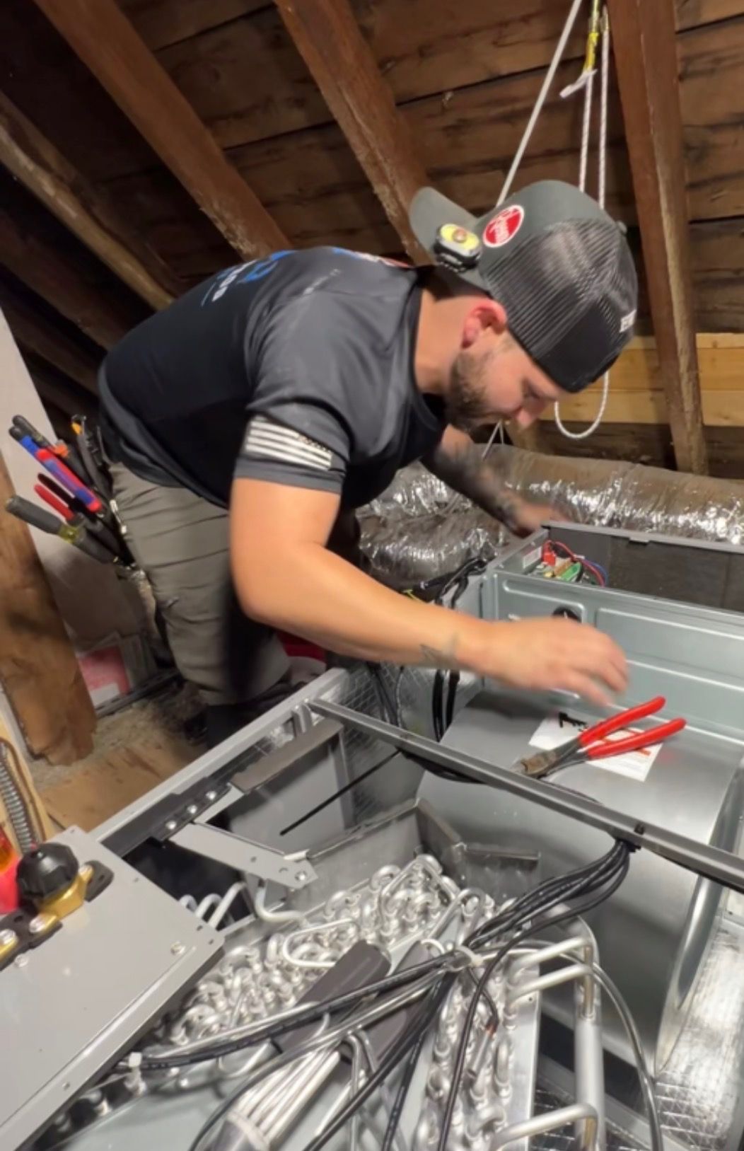 A person in a baseball cap works on an open HVAC unit in an attic, holding red-handled pliers.