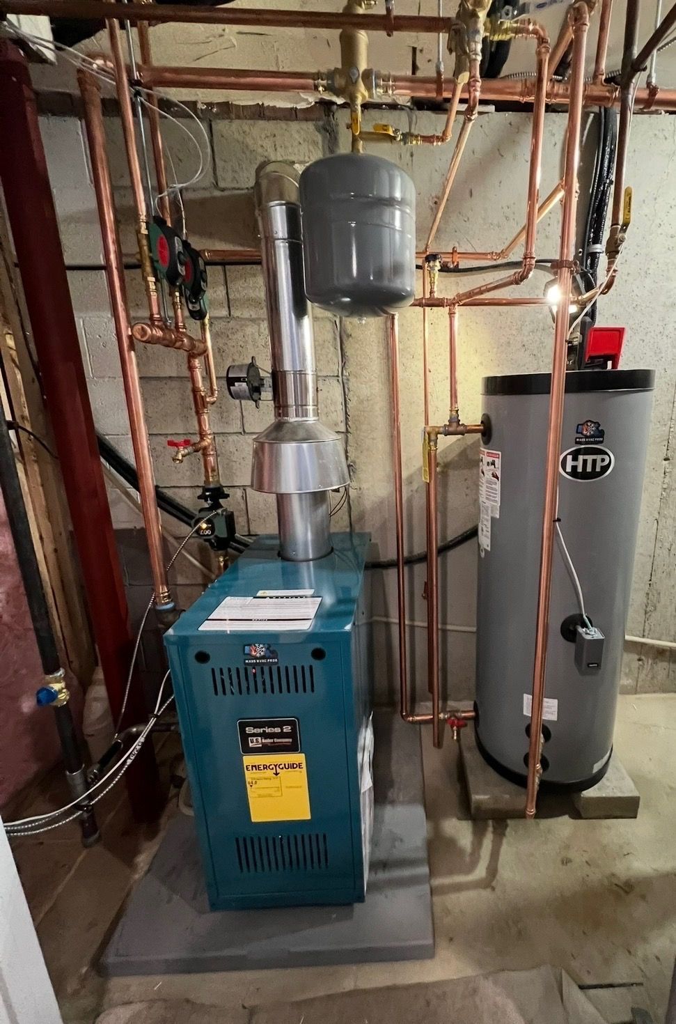 A blue boiler next to a gray HTP water tank with copper piping and an expansion tank in a basement.
