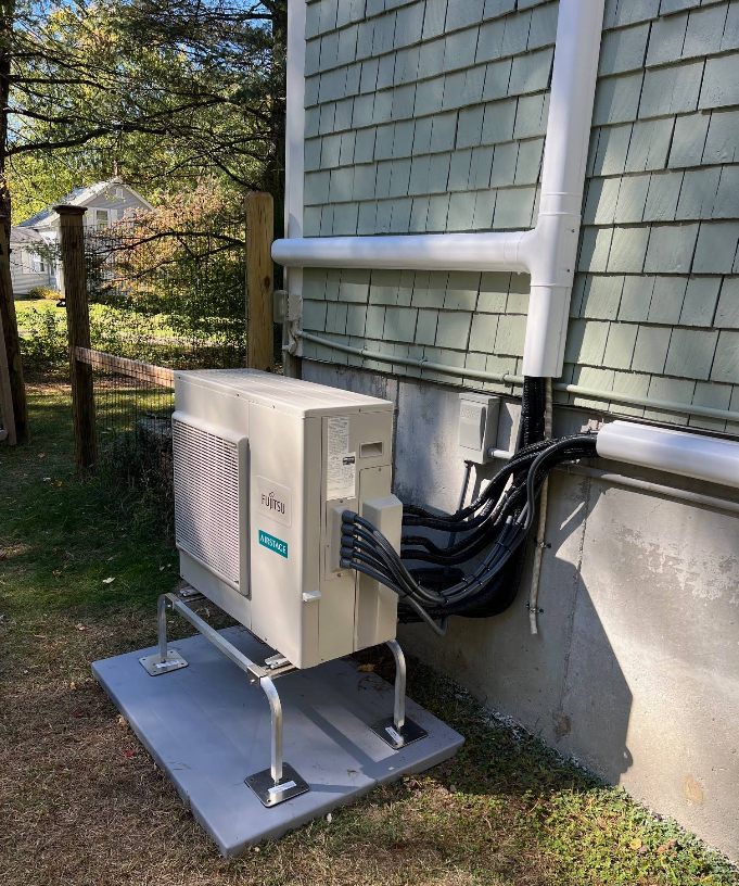 An outdoor heat pump unit sits on a mounting stand against a light green shingled house wall with protective conduit.