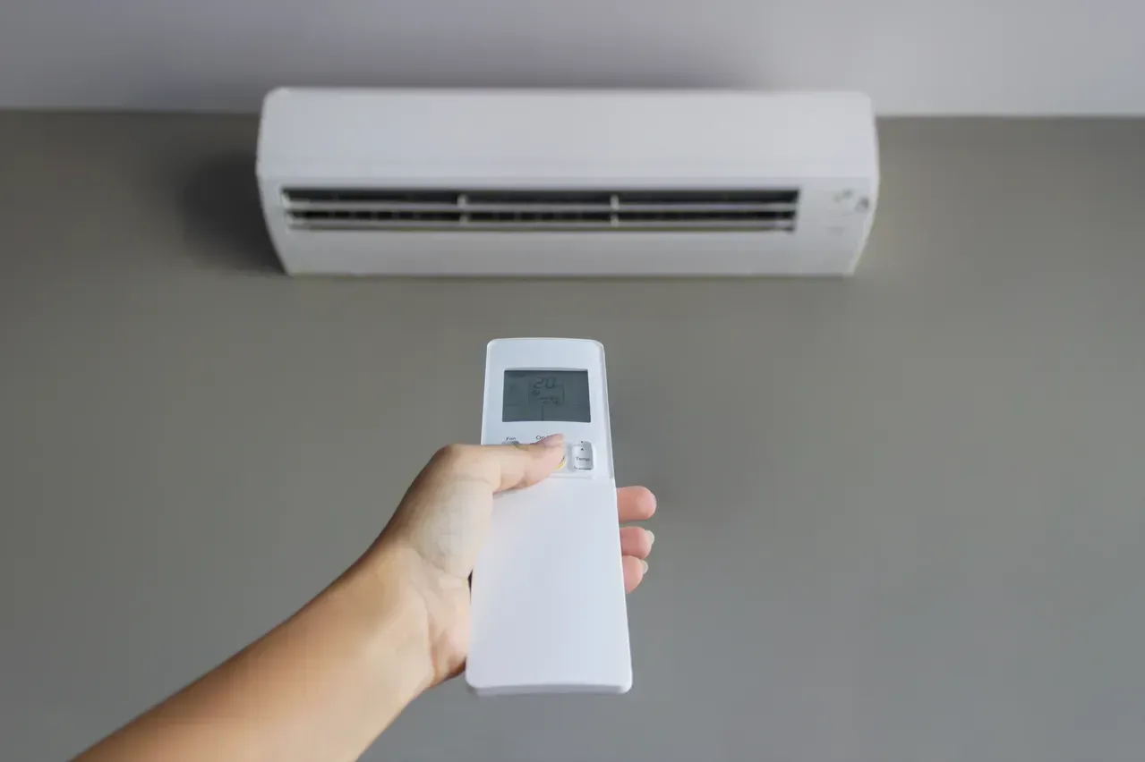 A hand holds a white remote control pointing towards a white wall-mounted air conditioner.