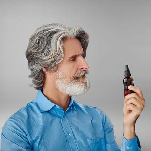 Une personne aux cheveux gris et à la barbe, vêtue d'une chemise bleue, tenant et observant un petit flacon compte-gouttes.