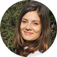 Une personne aux cheveux bruns mi-longs sourit doucement sur un fond flou de feuillage vert.