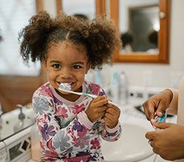 Un enfant sourit en se brossant les dents dans une salle de bain, avec une personne à ses côtés.