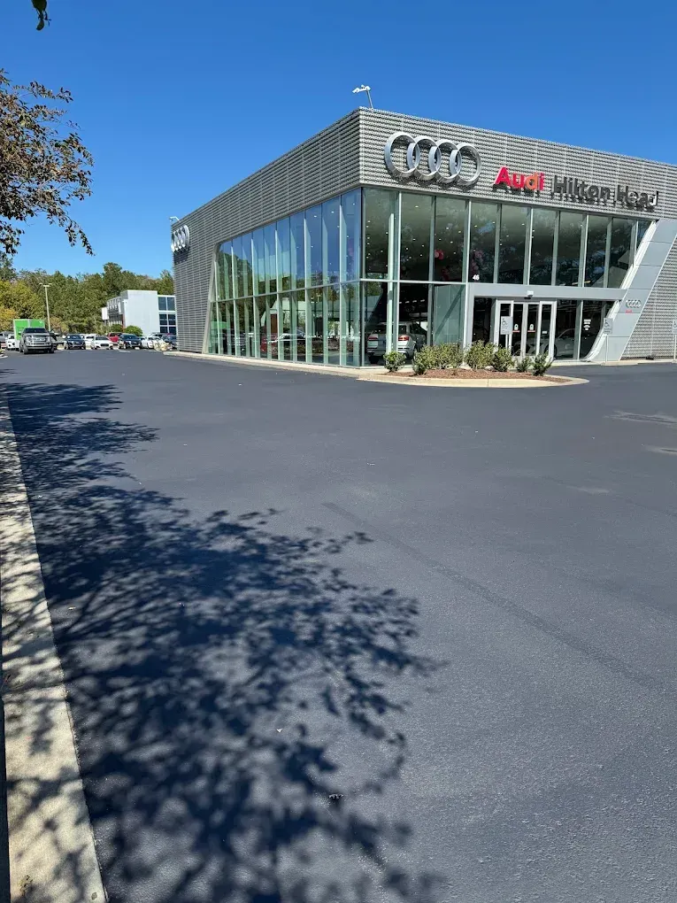 A modern glass and metal Audi dealership building seen from a wide, freshly paved parking lot under a clear blue sky.