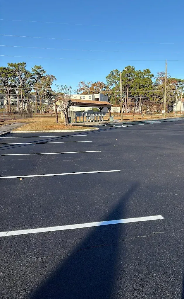 A paved parking lot with white painted lines leads toward a small, shaded outdoor seating area under a blue sky.