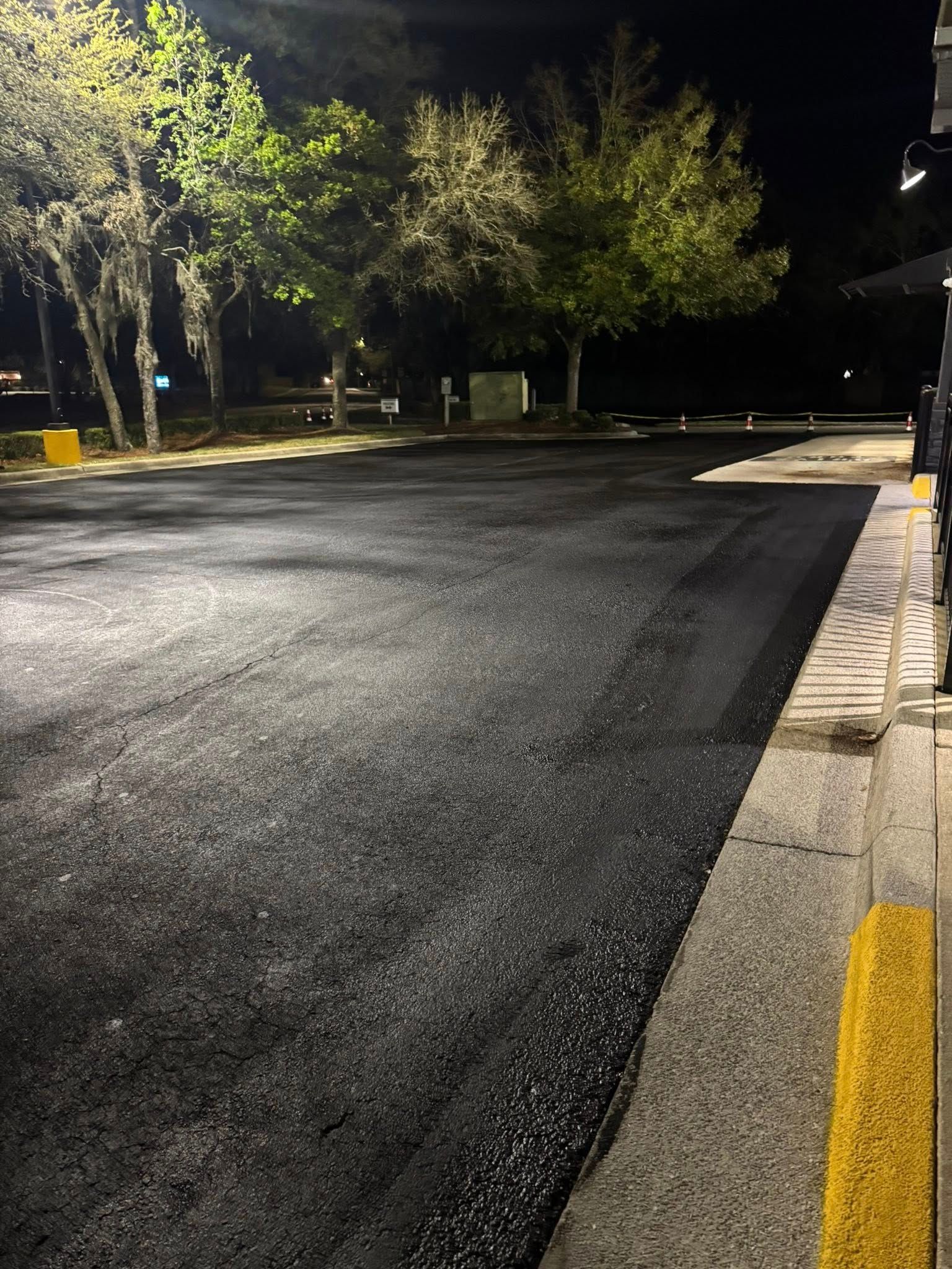 A freshly paved, dark asphalt parking lot at night, illuminated by overhead lights with trees in the background.