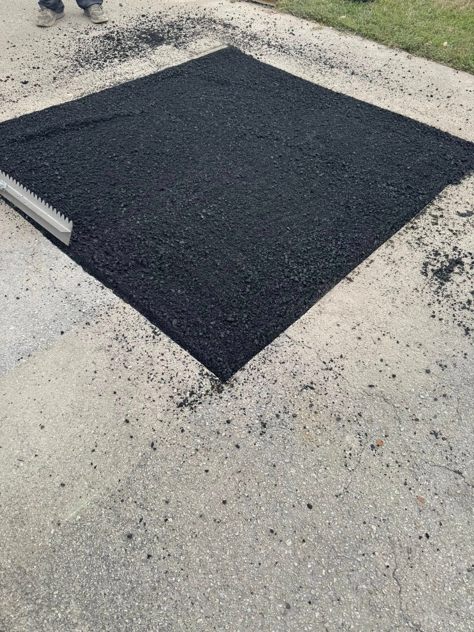 A fresh patch of black asphalt filling a square cut-out in a light gray concrete driveway.