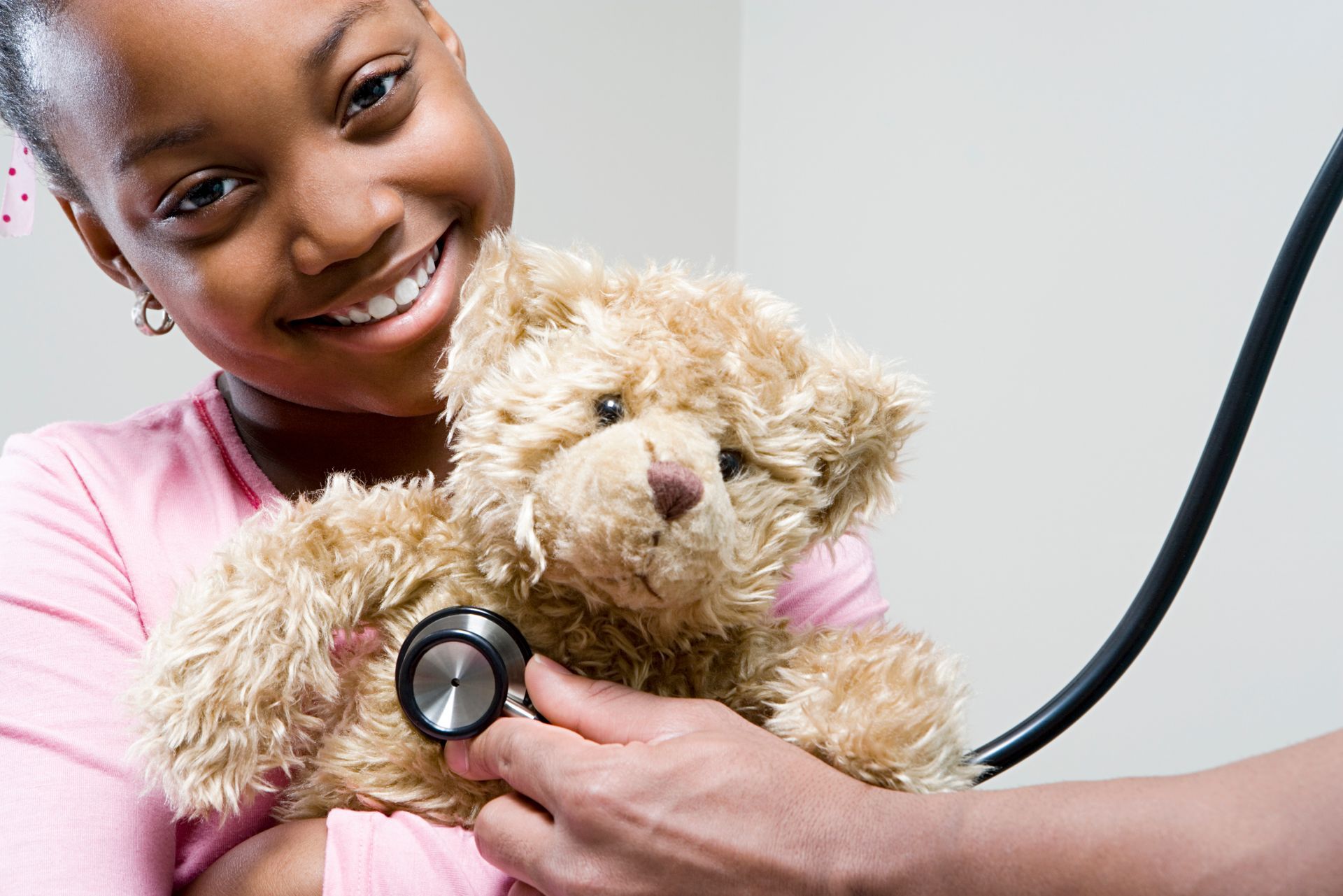 A smiling young girl is holding a teddy bear and a hand is placing a stethoscope on the teddy bear's chest.
