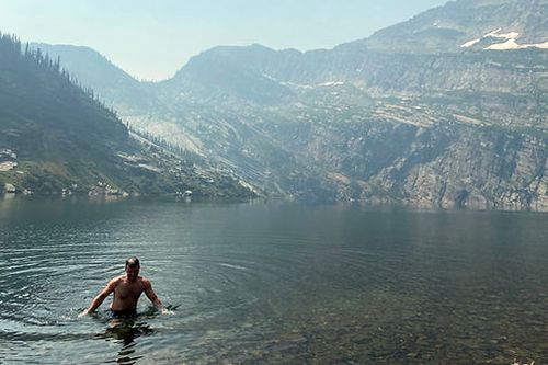 A man is swimming in a lake with mountains in the background.