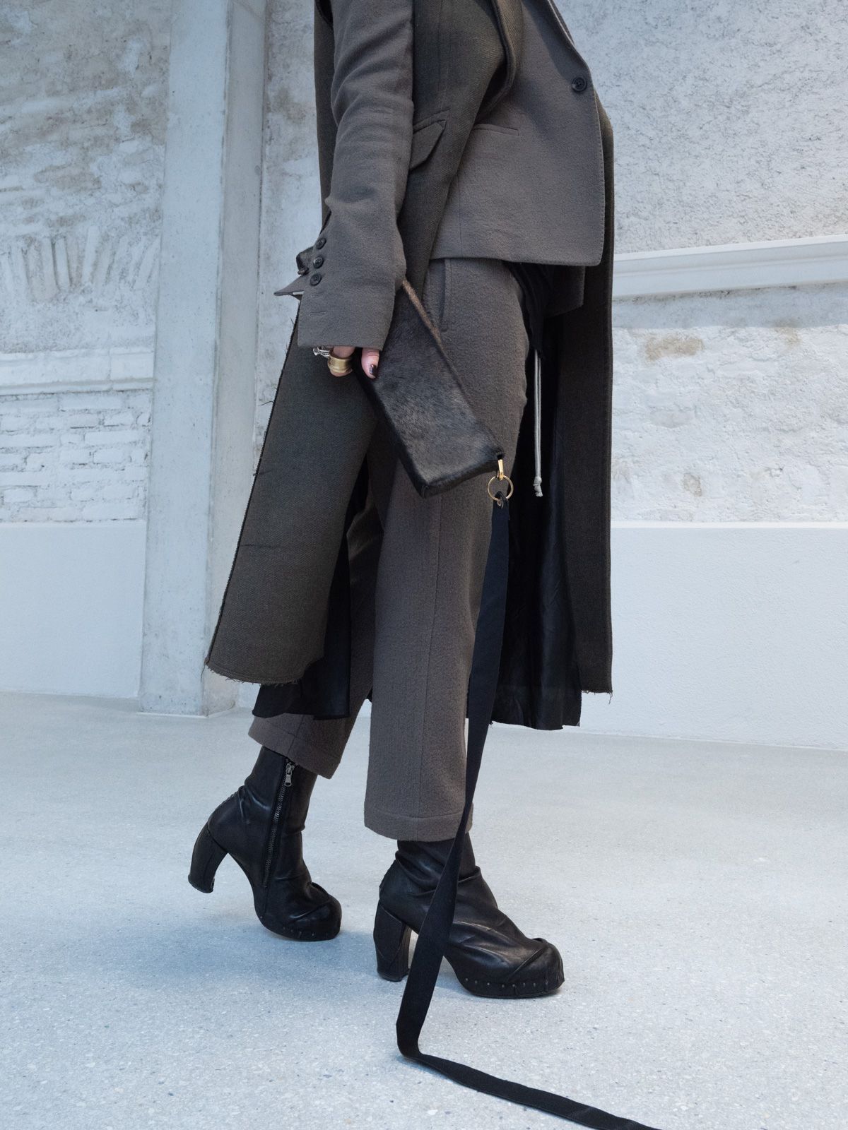 Eine Person in dunkler, mehrlagiger Kleidung geht und hält eine Clutch. Schwarze Stiefel, graue Hose und langer Mantel.