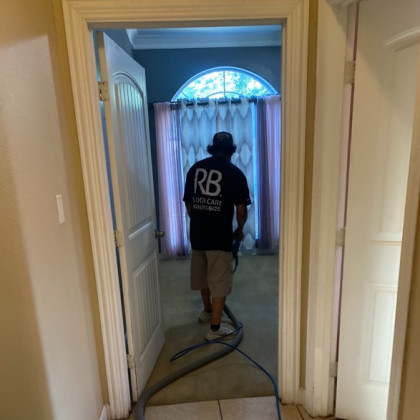 A person cleaning a carpet, seen through an open doorway. They are wearing a black shirt with a logo and khaki shorts.