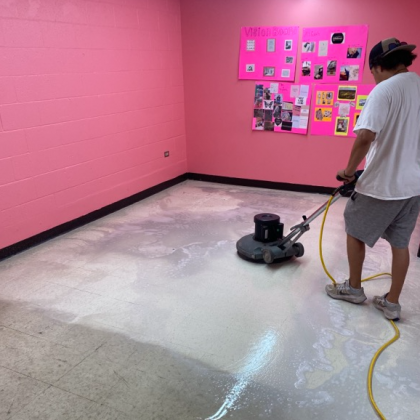 Man using floor buffer on a wet, light-colored floor in a pink-walled room.